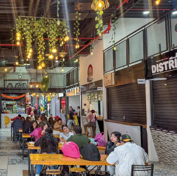 Plazoleta de comidas de VISTO llena de gente, con plantas y bombillos colgando sobre las mesas